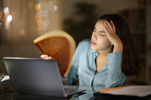 A woman fanning herself with a hand fan as she sits at a desk with a laptop.