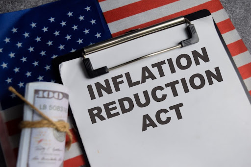 A clipboard with a paper that reads, "inflation reduction act." There's a roll of money nearby and an American flag in the background.